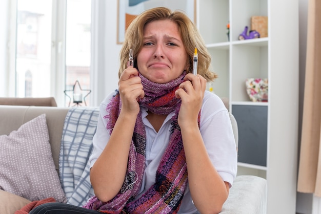 Jeune femme malsaine avec une écharpe chaude autour du cou, se sentant mal et malade souffrant de rhume et de grippe tenant une seringue et une ampoule ayant l'air inquiète et effrayée assise sur un canapé dans un salon clair