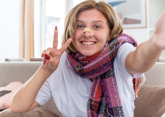 Jeune femme malsaine avec une écharpe chaude autour du cou souffrant de grippe et de rhume avec un patch sur le nez souriant montrant le signe v se sentant mieux confus assis sur un canapé dans un salon clair