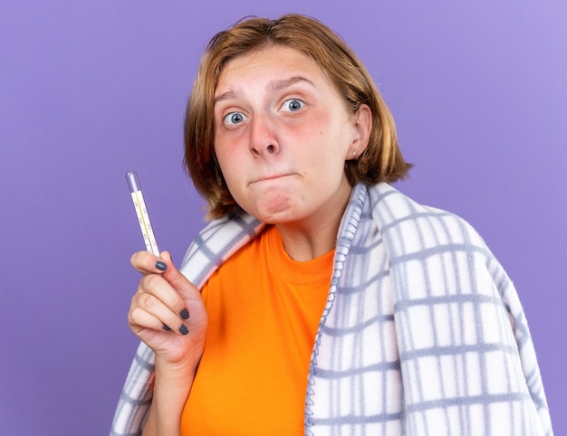 Jeune femme malsaine enveloppée dans une couverture chaude se sentant malade souffrant de grippe ayant de la fièvre mesurant sa température à l'aide d'un thermomètre l'air inquiète debout sur un mur violet