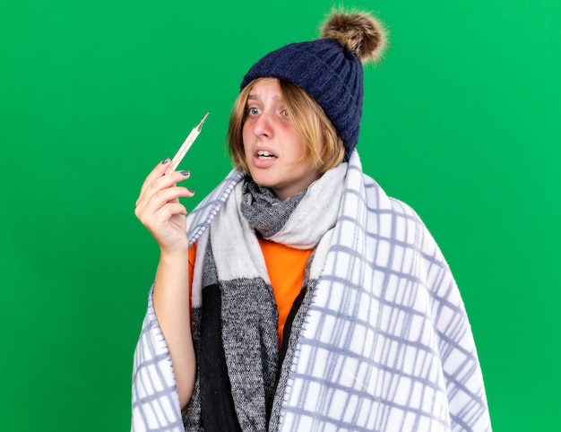 Jeune femme malsaine enveloppée dans une couverture portant un chapeau mesurant sa température corporelle à l'aide d'un thermomètre souffrant de grippe ayant de la fièvre l'air inquiet debout sur un mur vert