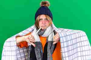 Photo gratuite jeune femme malsaine enveloppée dans une couverture portant un chapeau mesurant sa température corporelle à l'aide d'un thermomètre souffrant de grippe ayant de la fièvre montrant les pouces vers le bas debout sur un mur vert