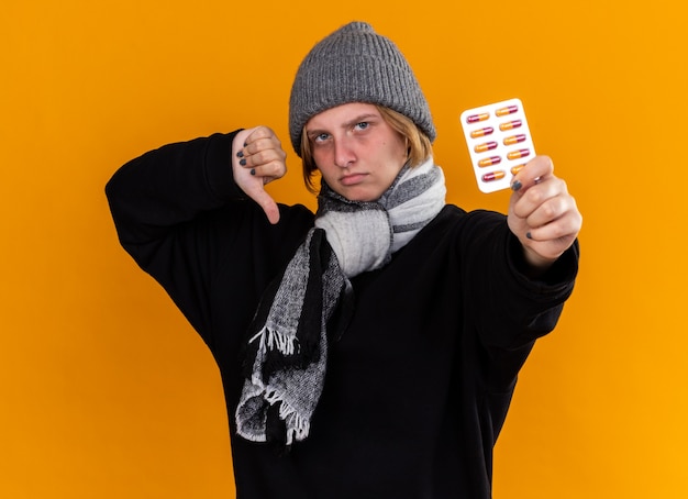 Photo gratuite jeune femme malsaine portant un chapeau chaud et une écharpe autour du cou se sentant malade souffrant de grippe tenant des pilules montrant les pouces vers le bas debout sur un mur orange