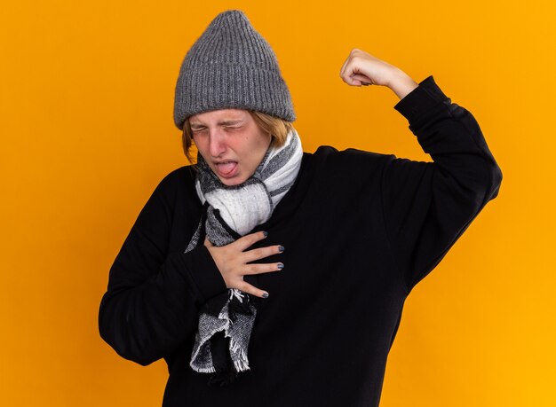 Jeune femme malsaine portant un chapeau chaud et une écharpe autour du cou se sentant malade souffrant de la grippe toussant debout sur un mur orange