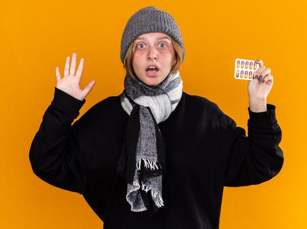 Jeune femme malsaine portant un chapeau chaud et une écharpe autour du cou se sentant terriblement souffrant du rhume et de la grippe tenant des pilules inquiètes debout sur un mur orange