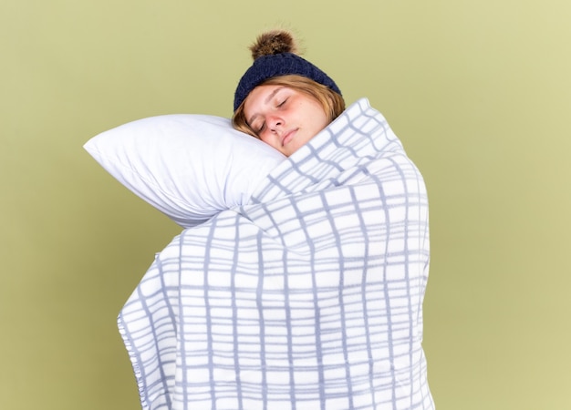 Une jeune femme malsaine portant un chapeau chaud enveloppé d'une couverture tenant un oreiller les yeux fermés veut dormir debout sur un mur vert