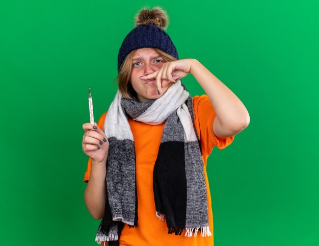 Jeune femme malsaine en t-shirt orange avec chapeau et écharpe chaude autour du cou se sentant terrible tenant un thermomètre ayant de la fièvre souffrant de nez qui coule debout sur un mur vert