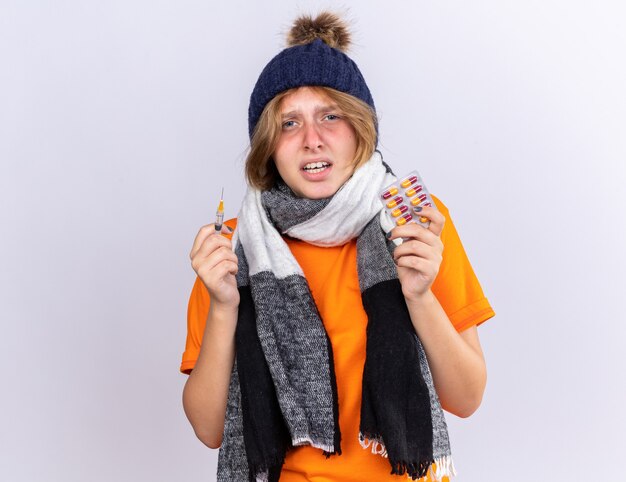 Jeune femme malsaine en t-shirt orange avec une écharpe chaude autour du cou et un chapeau se sentant terriblement souffrant de la grippe tenant une seringue et des pilules