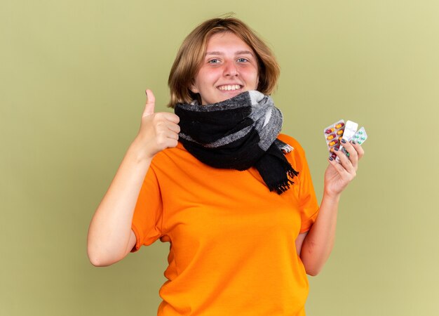 Jeune femme malsaine en t-shirt orange avec une écharpe chaude autour du cou se sentant mieux tenant différentes pilules souriantes montrant les pouces vers le haut debout sur le mur vert
