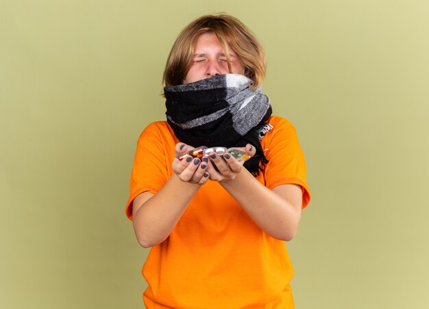 Jeune femme malsaine en t-shirt orange avec une écharpe chaude autour du cou se sentant terriblement souffrant de la grippe tenant différentes pilules semblant déçues debout sur un mur vert