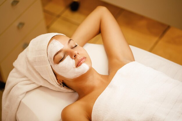 Jeune femme avec un masque facial blanc relaxant les yeux fermés au spa de beauté
