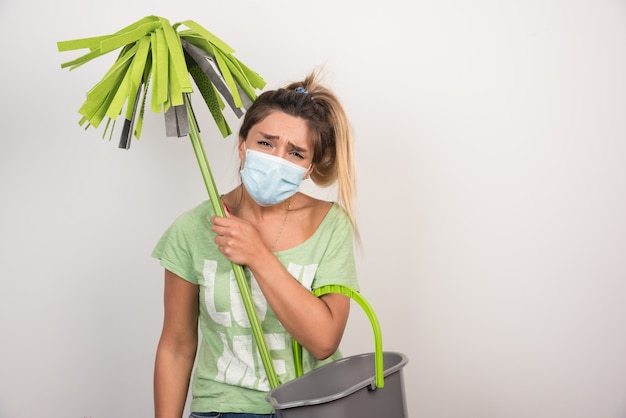 Jeune femme avec masque tenant une vadrouille sur un mur blanc.