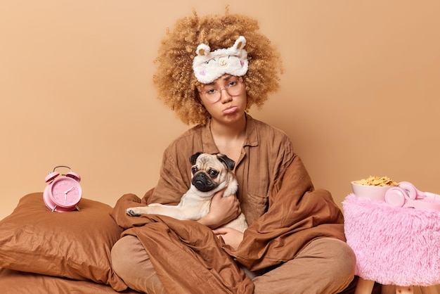 Une jeune femme mécontente aux cheveux bouclés enveloppée dans une couverture vêtue d'un costume de sommeil et d'un bandeau sur le front pose avec un chien carlin au lit a une mauvaise humeur se réveille tôt isolée sur fond marron
