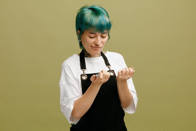 Une jeune femme mécontente portant un uniforme de coiffeur tenant un coupe-ongles regardant ses ongles isolés sur fond vert olive