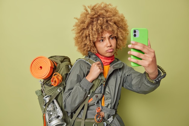 Une jeune femme mécontente sérieuse prend une vidéo d'enregistrements de selfie pour son blog de voyage se sent fatiguée prend une pause après avoir erré vêtue d'un coupe-vent porte un sac à dos isolé sur fond vert