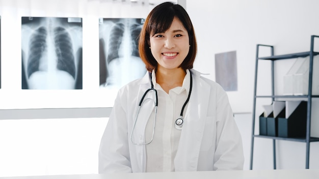 Jeune femme médecin asiatique confiante en uniforme médical blanc avec stéthoscope regardant la caméra et souriant lors d'une vidéoconférence avec un patient dans un hôpital de santé.