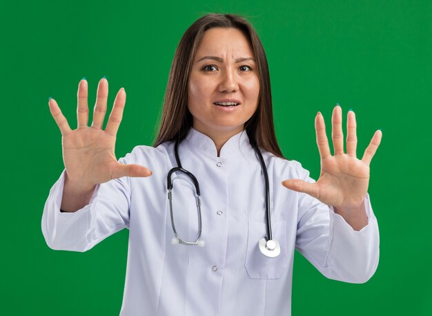 Jeune femme médecin asiatique effrayée portant une robe médicale et un stéthoscope regardant à l'avant faisant un geste de refus isolé sur un mur vert