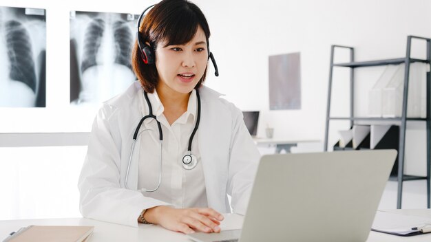 Jeune femme médecin asiatique en uniforme médical blanc avec stéthoscope utilisant un ordinateur portable parlant par vidéoconférence avec un patient au bureau dans une clinique de santé ou un hôpital.