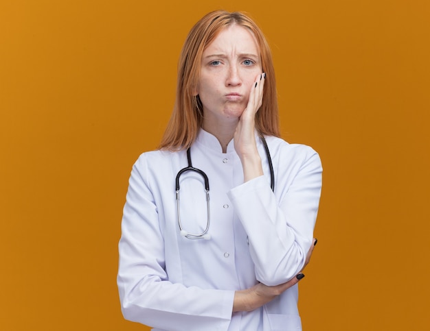 Jeune femme médecin au gingembre confuse portant une robe médicale et un stéthoscope touchant le visage