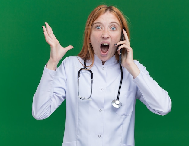 Photo gratuite jeune femme médecin au gingembre impressionnée portant une robe médicale et un stéthoscope parlant au téléphone regardant à l'avant montrant une main vide isolée sur un mur vert