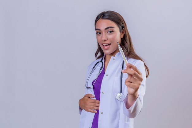 Jeune femme médecin en blouse blanche avec phonendoscope air confiant avec sourire sur le visage montrant un thermomètre sur fond blanc