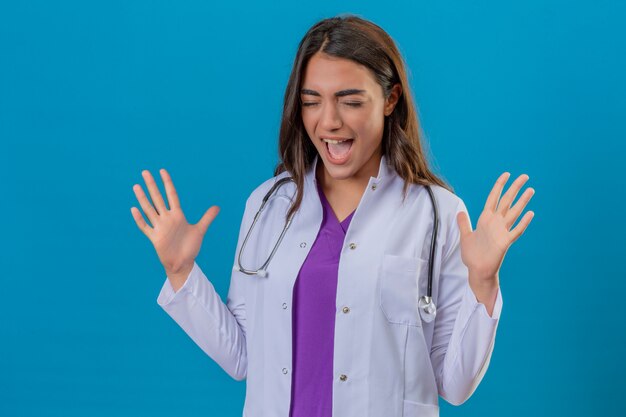 Jeune femme médecin en blouse blanche avec phonendoscope criant avec une expression agressive et les bras levés sur fond bleu isolé