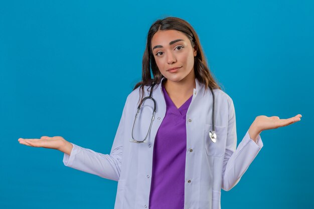 Jeune femme médecin en blouse blanche avec phonendoscope désemparé et expression confuse avec les bras et les mains levées sur fond bleu