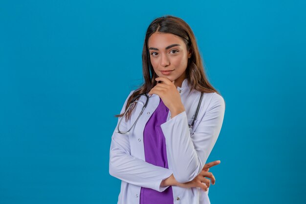 Jeune femme médecin en blouse blanche avec phonendoscope souriant menton heureux et confiant touchant avec la main sur fond isolé bleu