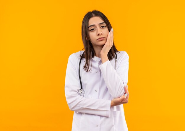 Jeune femme médecin en blouse blanche avec stéthoscope autour du cou à l'avant avec une expression pensive debout sur un mur orange