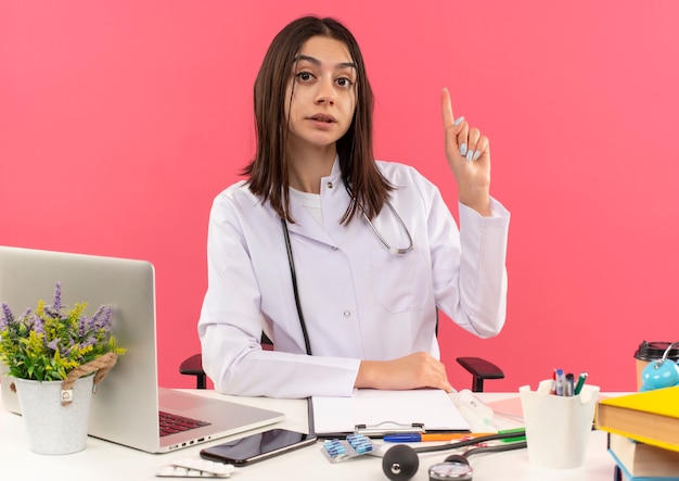 Jeune femme médecin en blouse blanche avec un stéthoscope autour du cou à l'avant montrant l'index avec un visage sérieux assis à la table avec un ordinateur portable sur un mur rose