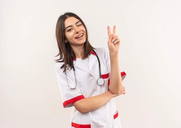 Jeune femme médecin en blouse blanche avec un stéthoscope autour du cou à l'avant en souriant et en clignant de l'œil montrant le signe de la victoire debout sur un mur blanc