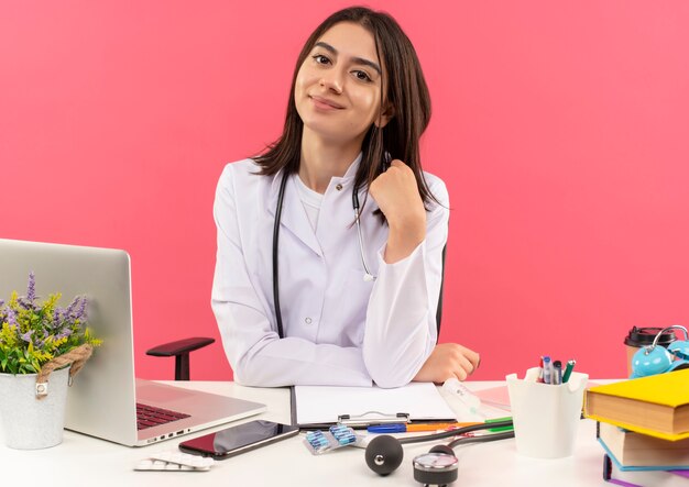 Jeune femme médecin en blouse blanche avec un stéthoscope autour du cou à l'avant souriant confiant assis à la table avec un ordinateur portable sur un mur rose