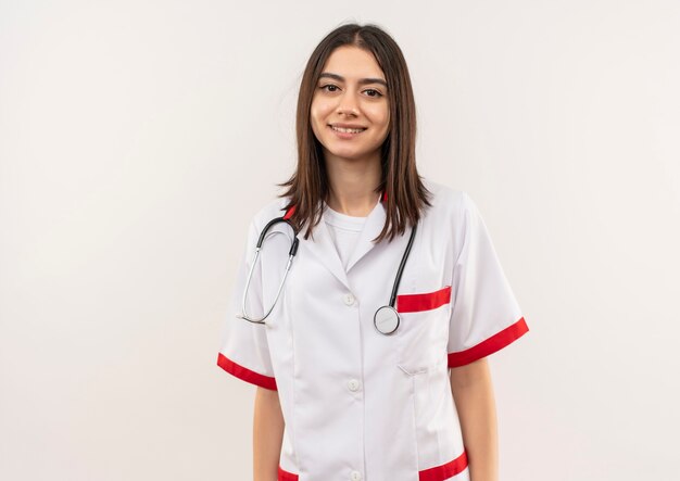 Jeune femme médecin en blouse blanche avec un stéthoscope autour du cou à l'avant souriant confiant debout sur un mur blanc