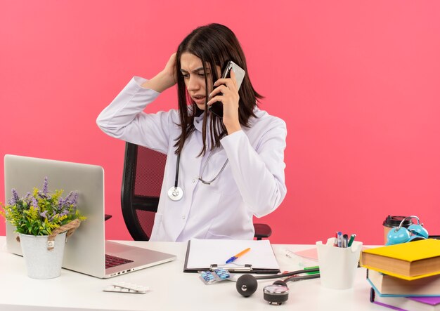 Jeune femme médecin en blouse blanche avec un stéthoscope autour du cou à la confusion tout en parlant au téléphone mobile assis à la table avec un ordinateur portable sur un mur rose