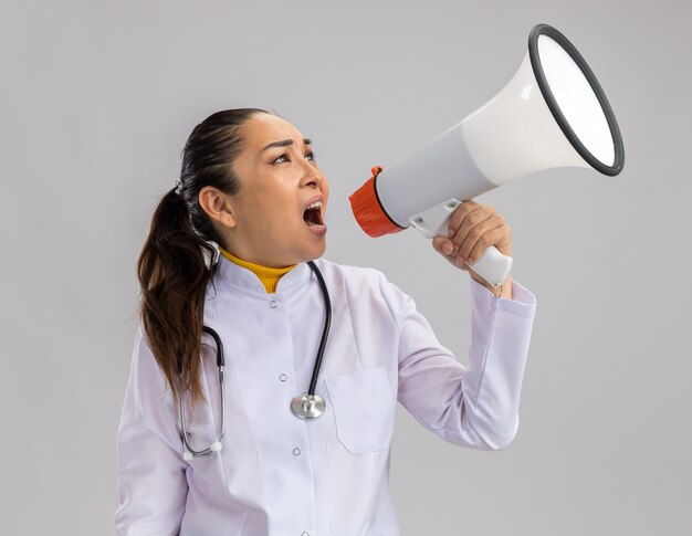 Jeune femme médecin en blouse blanche avec stéthoscope autour du cou criant au mégaphone avec une expression agressive debout sur un mur blanc