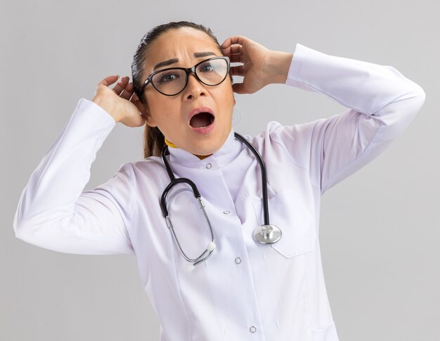 Jeune femme médecin en blouse blanche avec stéthoscope autour du cou criant avec une expression agacée touchant sa tête avec les mains