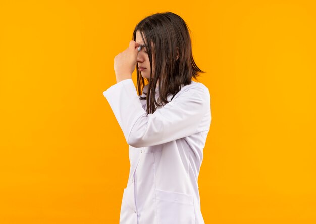 Jeune femme médecin en blouse blanche avec un stéthoscope autour du cou à la fatigue et surmené debout sur un mur orange