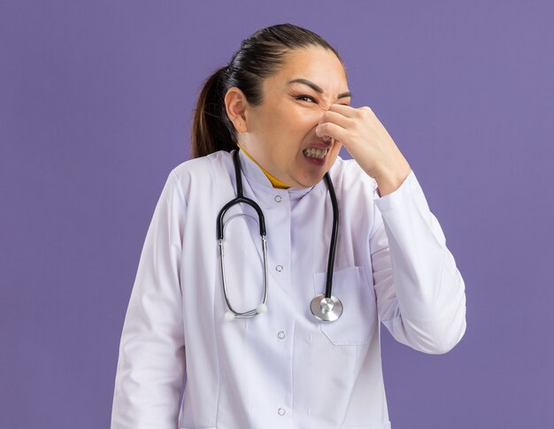 Jeune femme médecin en blouse blanche avec stéthoscope autour du cou fermant le nez avec des doigts souffrant de puanteur debout sur un mur violet