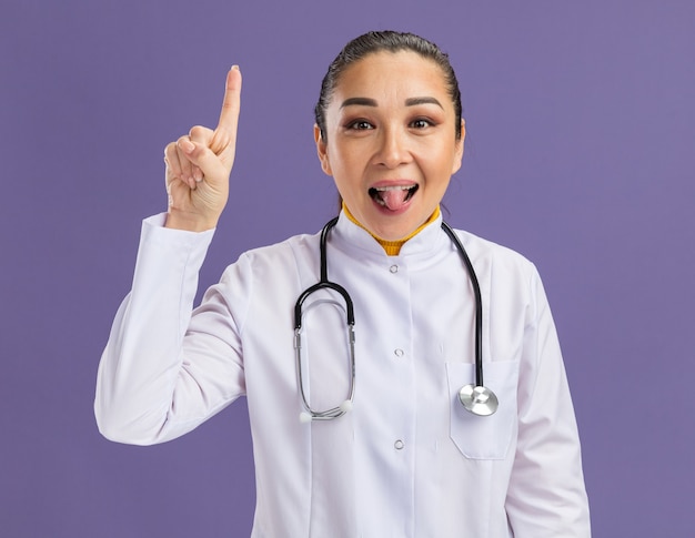 Jeune femme médecin en blouse blanche avec stéthoscope autour du cou heureux et surpris montrant l'index qui sort la langue debout sur le mur violet