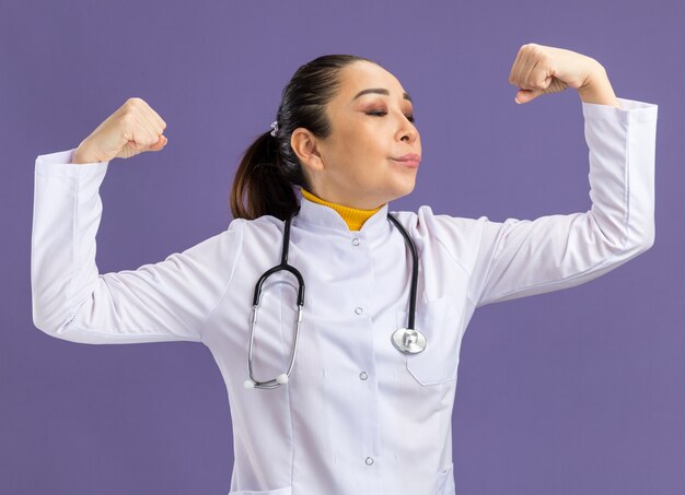 Jeune femme médecin en blouse blanche avec stéthoscope autour du cou levant les poings l'air confiant debout sur un mur violet