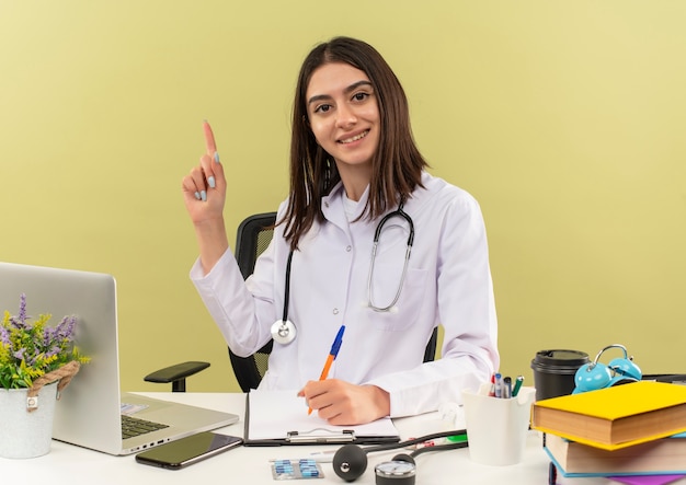 Jeune femme médecin en blouse blanche avec un stéthoscope autour du cou montrant l'index ayant une nouvelle idée assis à la table avec un ordinateur portable sur un mur léger