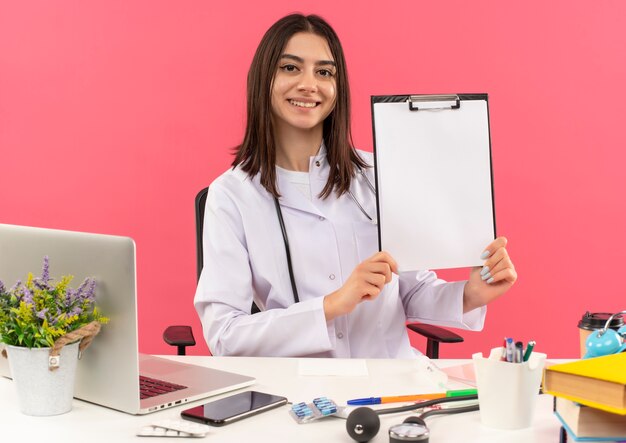 Jeune femme médecin en blouse blanche avec un stéthoscope autour du cou montrant le presse-papiers avec des pages vierges souriant à l'avant assis à la table avec un ordinateur portable sur un mur rose