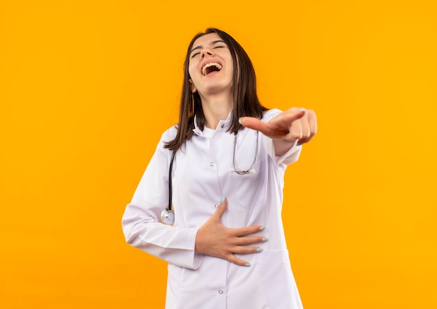 Jeune femme médecin en blouse blanche avec un stéthoscope autour du cou pointant avec le doigt pour vous rire debout sur un mur orange