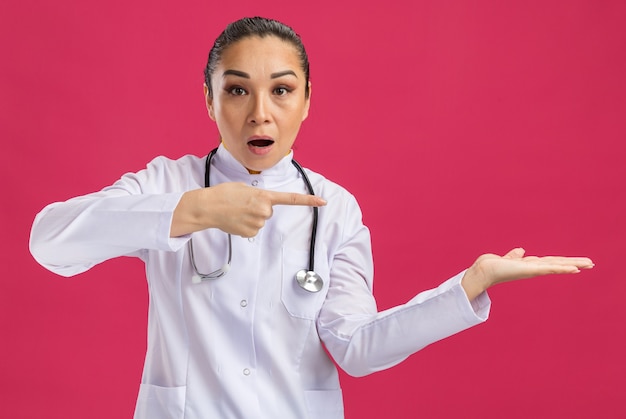 Jeune femme médecin en blouse blanche avec stéthoscope autour du cou présentant un espace de copie avec le bras pointé avec l'index debout sur le mur rose