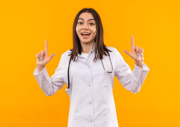 Jeune femme médecin en blouse blanche avec un stéthoscope autour du cou à la recherche à l'avant souriant heureux et positif montrant joyeusement l'index debout sur le mur orange