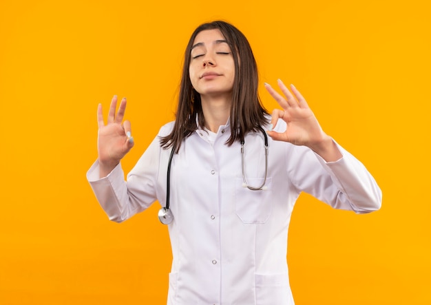 Jeune femme médecin en blouse blanche avec stéthoscope autour du cou se détendre avec les yeux fermés faisant le geste de méditation avec les doigts debout sur le mur orange