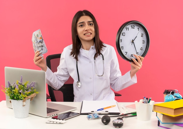 Jeune femme médecin en blouse blanche avec un stéthoscope autour du cou tenant de l'argent et une horloge murale à la confusion et très anxieux assis à la table avec un ordinateur portable sur un mur rose