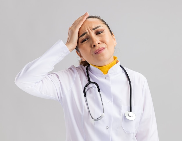 Jeune femme médecin en blouse blanche avec stéthoscope autour du cou touchant sa tête avec une expression agacée debout sur un mur blanc