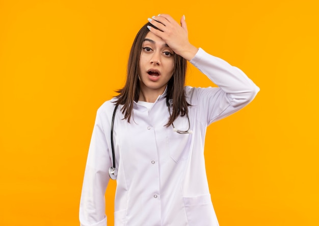 Jeune femme médecin en blouse blanche avec un stéthoscope autour de son cou à l'avant confus, oublié, mauvaise mémoire concept debout sur un mur orange
