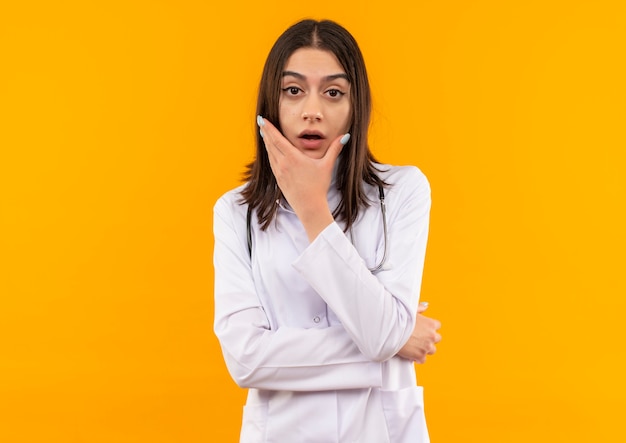 Jeune femme médecin en blouse blanche avec un stéthoscope autour de son cou à l'avant avec une expression pensive perplexe debout sur un mur orange