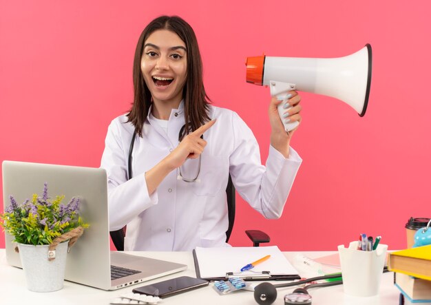 Jeune femme médecin en blouse blanche avec un stéthoscope autour de son cou tenant un mégaphone pointant avec le doigt vers lui souriant joyeusement assis à la table avec un ordinateur portable sur un mur rose